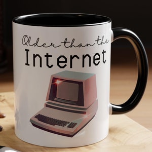 Puede incluir: Taza de cerámica blanca con borde y asa negros. La taza presenta un gráfico de computadora retro y el texto "Older than the Internet".