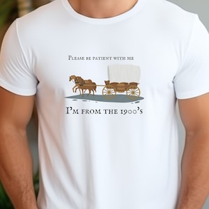 May include: White t-shirt with a brown illustration of a covered wagon and horses. The text says, "Please be patient with me, I'm from the 1900's."