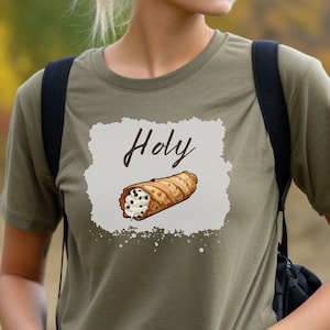 May include: Olive green t-shirt featuring a cartoon cannoli with the word "Holy" above it. The cannoli is brown with white cream and chocolate chips. The text and graphic are on a light gray background.
