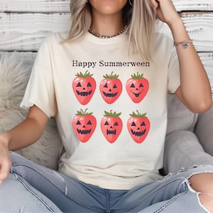 May include: A cream-coloured t-shirt featuring the words "Happy Summerween" above six red strawberry jack-o'-lanterns. The strawberries have green stems and carved faces. A casual, seasonal design.