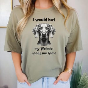 Op de afbeelding: Olijfgroen t-shirt met een zwart-wit illustratie van een Weimaraner hond. De tekst "I would but my Weimie needs me home" staat boven en onder de afbeelding van de hond.