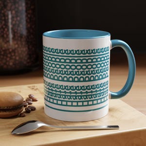 May include: White ceramic coffee mug with a teal interior, handle, and decorative teal geometric pattern. The mug sits on a wooden cutting board with a spoon and a macaron.
