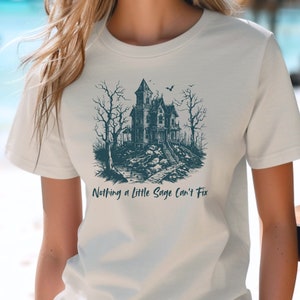 May include: A white t-shirt with a dark blue illustration of a spooky house with a bat flying overhead. The text "Nothing a little Sage Can't Fix" is printed below the illustration.