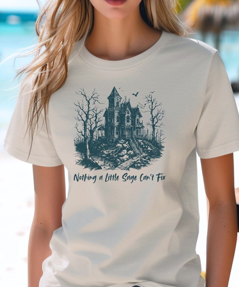 May include: A white t-shirt with a dark blue illustration of a spooky house with a bat flying overhead. The text "Nothing a little Sage Can't Fix" is printed below the illustration.