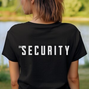 Op de afbeelding: Zwart T-shirt met het woord "SECURITY" in grote, witte blokletters op de rug. Het woord "IN" is in kleinere witte letters voor het woord "SECURITY" gedrukt. Het shirt is gemaakt van een zacht materiaal.