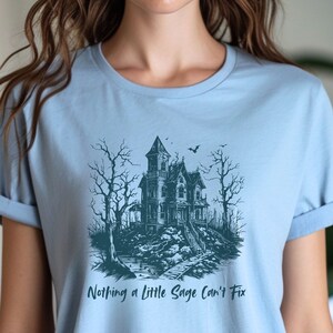 May include: Light blue t-shirt with a graphic of a spooky house in a forest with the text "Nothing a little Sage can't fix".