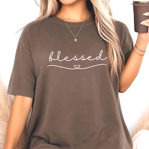 以下が含まれることがあります： 前面に白い筆記体で「blessed」という文字が書かれた茶色の半袖Tシャツ。単色のシャツで、ゆったりとしたフィット感です。単語の下には小さなハートが描かれています。