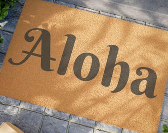 May include: A rectangular doormat with the word "Aloha" in dark gray script. The mat is a light brown color and sits on a gray stone surface. Green plants are visible in the background.