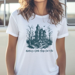 May include: White t-shirt with a dark blue illustration of a spooky house with a bat flying overhead. The text "Nothing a little Sage can't fix" is printed below the illustration.
