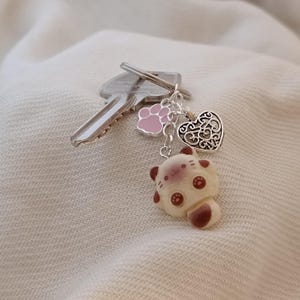 Può includere: Un portachiavi con un ciondolo a forma di gatto color crema con macchie marroni e una piccola coda, un ciondolo a forma di zampa rosa e un ciondolo a forma di cuore argentato. Due chiavi sono attaccate al portachiavi.