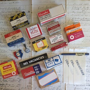 May include: A collection of vintage office supplies, including boxes of staples, paper clips, and reinforcements, along with a pen and labels, arranged on a ledger sheet. The items feature various brand names and colorful packaging.