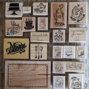 May include: A collection of wooden rubber stamps featuring various kitchen and food-themed designs. The stamps include illustrations of cakes, coffee, tea, utensils, and recipe cards with text such as You Take The Cake, Congratulatte, and Menu.