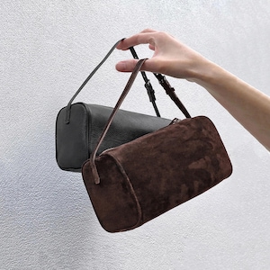 May include: Two cylindrical handbags with adjustable straps. One bag is made of black textured leather, and the other is made of dark brown suede. Both bags feature a rounded, tube-like shape and are suspended by thin, matching shoulder straps.