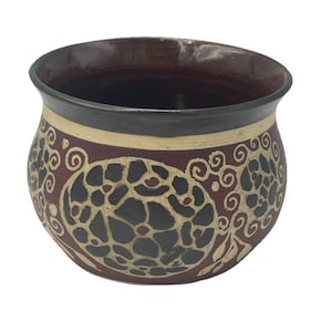 May include: A handcrafted ceramic bowl with a dark brown interior and rim. The exterior features a maroon base with a cream-colored band and intricate black and cream-colored swirling patterns. A unique decorative piece.