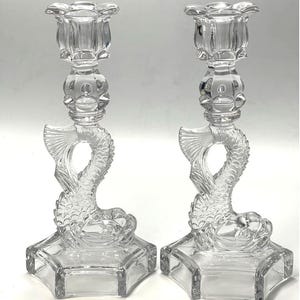 May include: Two clear glass candle holders. Each holder features a fish design with a detailed tail and body. The candle holders have a hexagonal base and a floral-shaped top. The glass is transparent, allowing light to pass through.