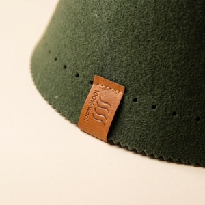 May include: A dark green felt cone with a textured surface and a row of small perforations along the bottom edge. A tan leather tag is stitched to the side, embossed with the text 100% wool and a graphic icon of three wavy steam lines.