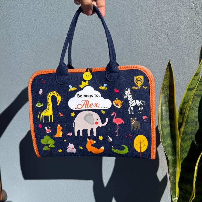 May include: A navy blue tote bag with orange trim and handles. The bag features colorful illustrations of animals, including a giraffe, elephant, and zebra. The text "Belongs to: Alex" is displayed on the front.