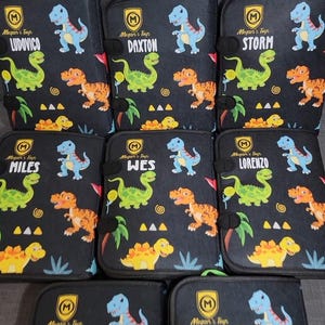 May include: A collection of black rectangular cases featuring a repeating dinosaur pattern in blue, green, and orange. Each case displays a yellow shield logo with the letter M, the text Megan's Toys, and a unique name: Ludovico, Daxton, Storm, Miles, Wes, or Lorenzo.