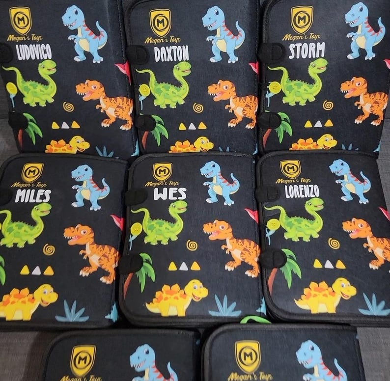 May include: A collection of black rectangular cases featuring a repeating dinosaur pattern in blue, green, and orange. Each case displays a yellow shield logo with the letter M, the text Megan's Toys, and a unique name: Ludovico, Daxton, Storm, Miles, Wes, or Lorenzo.