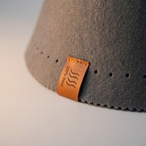May include: A dark gray felt cone with a textured surface and a row of small, evenly spaced perforations along the bottom edge. A tan leather tag is stitched onto the felt, featuring the embossed text 100% Wool and a wavy line icon.