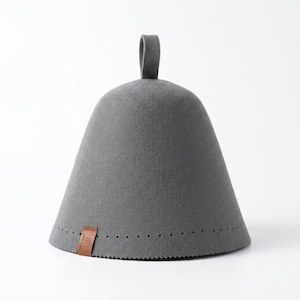 May include: A conical grey felt sauna hat with a small loop at the top. The base features a row of small perforations and a brown leather tag with the text 100% Wool and a wavy line icon. The hat has a smooth, dense texture throughout.