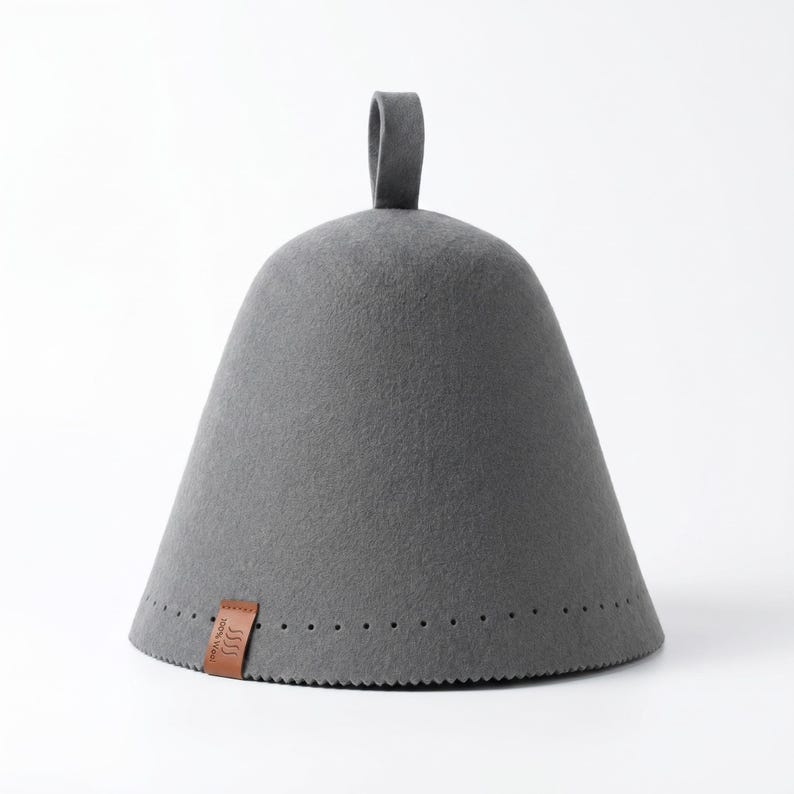 May include: A conical grey felt sauna hat with a small loop at the top. The base features a row of small perforations and a brown leather tag with the text 100% Wool and a wavy line icon. The hat has a smooth, dense texture throughout.