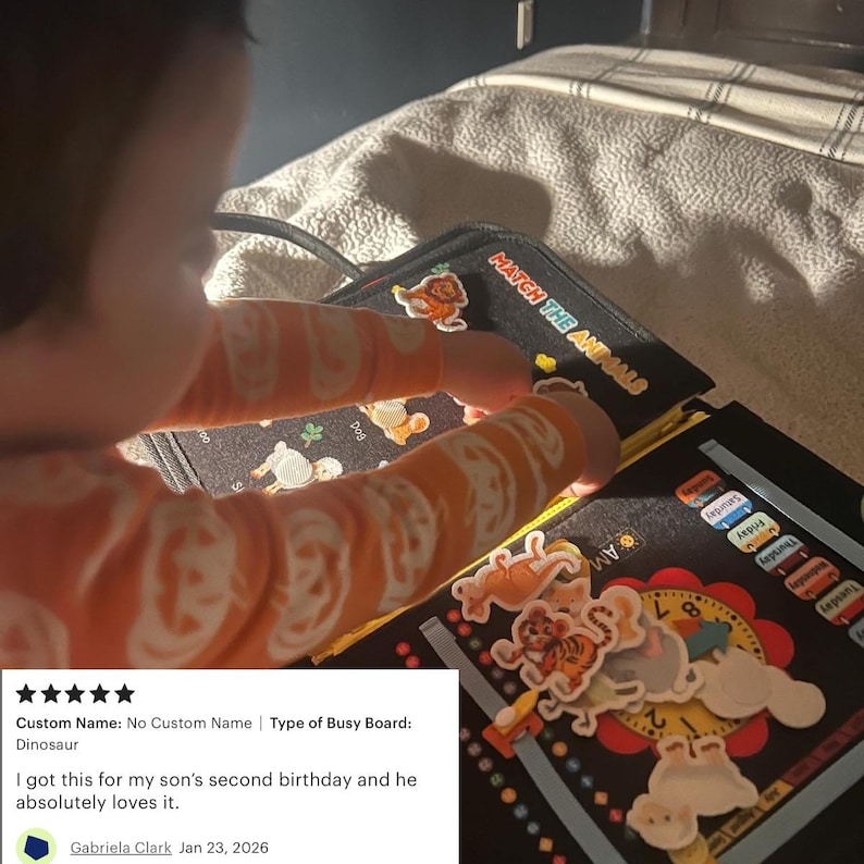 May include: A black busy board with animal-themed illustrations and the text "MATCH THE ANIMALS." The board features colorful animal cutouts and a clock design. A child's hands are interacting with the board.