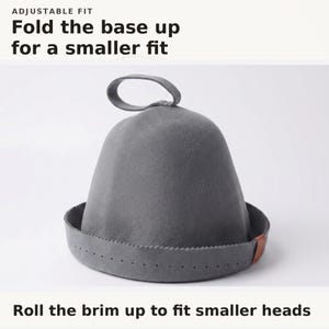 May include: A grey felt hat with a loop at the top and a folded brim featuring a small brown leather accent. Text at the top reads ADJUSTABLE FIT Fold the base up for a smaller fit. Text at the bottom reads Roll the brim up to fit smaller heads.