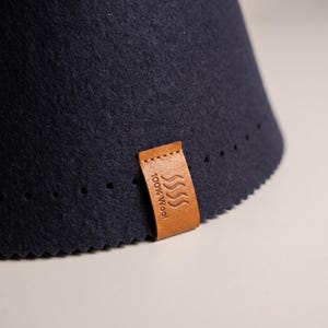 May include: A dark navy blue felted wool cone-shaped item with a zigzag cut bottom edge. A tan leather tag is attached to the side, featuring an embossed wavy line icon and the text 100% Wool. A row of small circular perforations runs horizontally.