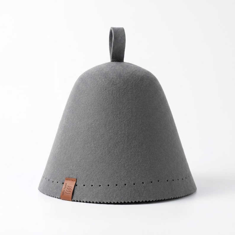 May include: A conical grey felt sauna hat with a small loop at the top. The base features a row of small perforations and a brown leather tag with the text 100% Wool and a wavy line icon. The hat has a smooth, dense texture throughout.