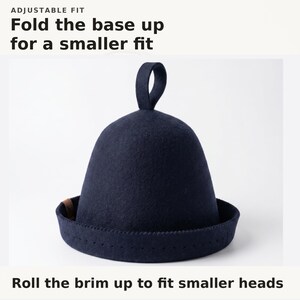 May include: A dark navy blue felt hat with a rounded crown, a small loop at the top, and a wide brim. The text above reads ADJUSTABLE FIT Fold the base up for a smaller fit, and the text below reads Roll the brim up to fit smaller heads.