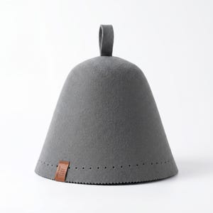 May include: A conical grey felt sauna hat with a small loop at the top. The base features a row of small perforations and a brown leather tag with the text 100% Wool and a wavy line icon. The hat has a smooth, dense texture throughout.