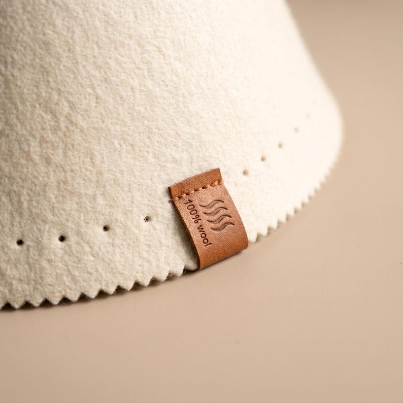 May include: A cream-coloured felted wool cone with a zigzag-cut bottom edge and a row of small perforations. A brown leather tag is stitched to the side, embossed with a wavy line icon and the text 100% wool in a clear, sans-serif font.