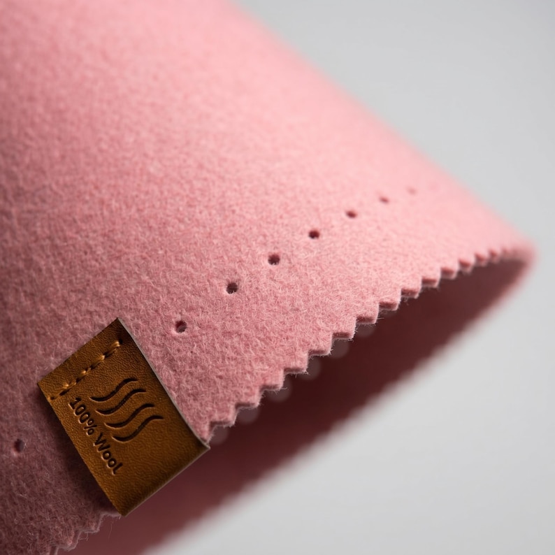 May include: A piece of soft pink felt fabric with a zigzag cut edge and a row of small perforations. A rectangular brown leather tag is stitched to the corner, featuring a wavy line icon and the printed text 100% Wool in black ink.