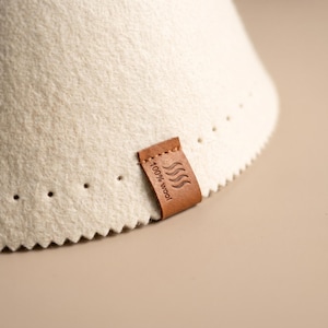 May include: A cream-coloured felted wool cone with a zigzag-cut bottom edge and a row of small perforations. A brown leather tag is stitched to the side, embossed with a wavy line icon and the text 100% wool in a clear, sans-serif font.