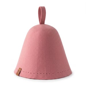 May include: A dusty rose felt sauna hat with a conical shape and a top hanging loop. The base features a decorative row of small perforations and a small brown rectangular leather tag attached to the side with embossed text on the surface.