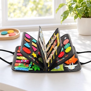May include: A black felt activity book featuring various interactive elements including colorful geometric shapes, buttons, buckles, and straps. The pages display text labels such as My Shapes and My Numbers, with assorted tactile components attached.