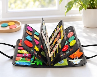 May include: A black felt activity book featuring various interactive elements including colorful geometric shapes, buttons, buckles, and straps. The pages display text labels such as My Shapes and My Numbers, with assorted tactile components attached.