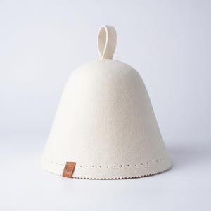 May include: A conical sauna hat made of off-white felted material. The hat features a small loop at the top for hanging and a brown rectangular leather tag near the bottom edge, which is decorated with a row of small, evenly spaced perforations.