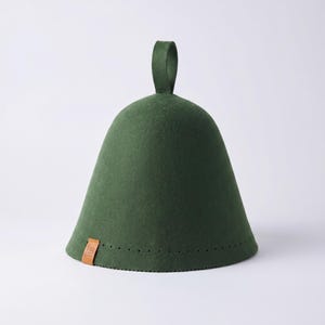 May include: A dark green felt sauna hat with a bell-shaped silhouette and a vertical loop handle at the top. The base features a row of small perforations and a small, rectangular tan leather tag attached to the lower edge of the hat.