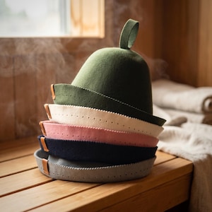 May include: A stack of five felt sauna hats in olive green, cream, dusty pink, navy blue, and light grey. Each hat features a rounded crown, a small top loop, a wide brim with perforated edges, and a small brown leather tab on the side.