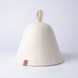 May include: A conical sauna hat made of off-white felted material. The hat features a small loop at the top for hanging and a brown rectangular leather tag near the bottom edge, which is decorated with a row of small, evenly spaced perforations.