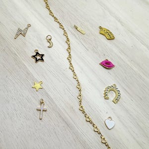 May include: A gold chain bracelet with a heart-shaped link design. The bracelet is shown with a variety of charms, including a star, a crescent moon, a lightning bolt, a cross, a horseshoe, a playing card, a heart, and a pair of lips.