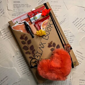 May include: A brown paper package with the words "BINX BOOKS" written on it. The package contains a notebook, a pen, stickers, a fluffy heart-shaped keychain, and other small items. The package is surrounded by pages of a book with text about love, self-love, and forgiveness.