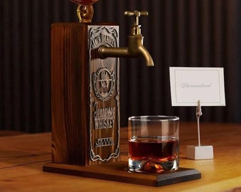 Op de afbeelding: Een houten whiskydispenser met een messing kraan en een glas amberkleurige vloeistof. De dispenser is voorzien van het logo "Jack Daniel's Tennessee Whiskey". Een kleine kaart met het woord "Gepersonaliseerd" wordt weergegeven.