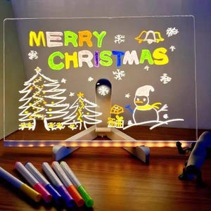 May include: A clear, illuminated Christmas-themed message board with the words "MERRY CHRISTMAS" in colorful letters. The board features drawings of Christmas trees, a snowman, and snowflakes. Includes a set of colorful markers.