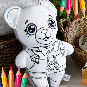 Op de afbeelding: Een witte teddybeer met zwarte omtrekdetails, waaronder een vlinderdas en een tuinbroek. De beer heeft grote ogen en een lachend gezicht. Kleurpotloden zijn rond de beer gerangschikt, wat duidt op een kleuractiviteit.
