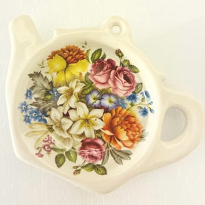 Könnte beinhalten: Ein cremefarbener Teebeutelhalter aus Keramik in Teekannenform mit einem zentralen Blumenarrangement aus gelben, orangefarbenen, rosafarbenen und weißen Blüten, grünen Blättern und kleinen blauen Blumen sowie einem Loch zum Aufhängen.