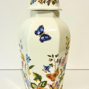 May include: A white porcelain ginger jar with a matching lid, featuring a colorful botanical pattern of various flowers and butterflies. The hexagonal vessel displays blue, yellow, orange, and pink floral motifs alongside detailed butterflies.