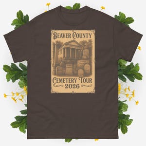 May include: A brown t-shirt featuring a vintage-style graphic for the "Beaver County Cemetery Tour 2026." The design includes an illustration of a classical building and tombstones, with text in a decorative font.